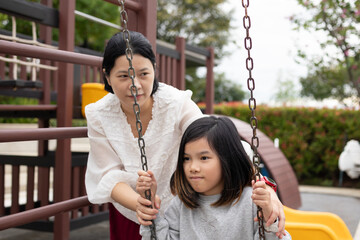 Happy Asian mother gently pushing her daughter on a swing in the outdoor playground, enjoying active family bonding time