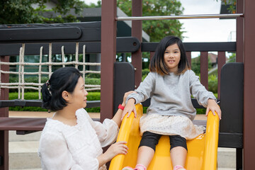 Young Asian girl sitting on yellow slide in playground while her mother supervises from behind, enjoying outdoor family fun