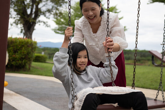 Happy Asian mother gently pushing her daughter on a swing in the outdoor playground, enjoying active family bonding time - Powered by Adobe