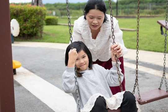 Happy Asian mother gently pushing her daughter on a swing in the outdoor playground, enjoying active family bonding time - Powered by Adobe