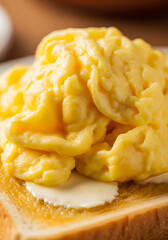 A vertical close-up of a generous pile of soft, fluffy, creamy scrambled eggs resting on a slice of golden-brown buttered toast.