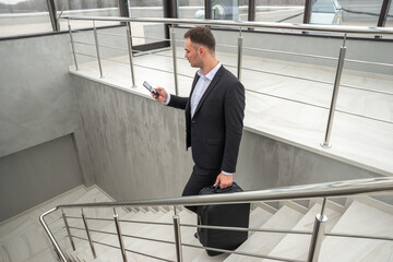 Young businessman with suitcase in stylish suit talking by phone while walking by stairs in modern business building