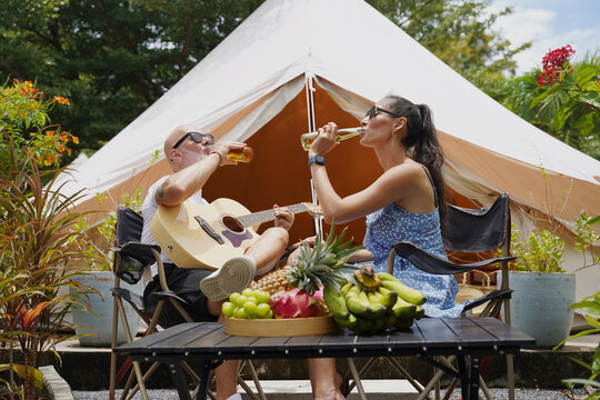A man and woman is joyfully drinking beer and playing guitar near a cozy tent at glamping