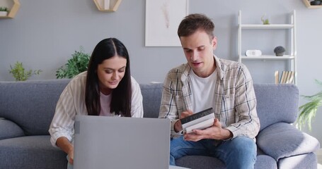 Happy young couple sitting on the couch, using a laptop and a calculator, paying house bills, tracking monthly expenses, managing family finances and planning budget together - Powered by Adobe