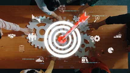 Aerial view of diverse team members collaborating over a table with gears and a glowing target,...