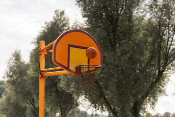 From above the rim basketball finds its mark in the orange and yellow hoop, set against a backdrop of leafy green trees