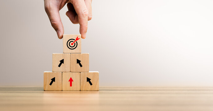 Business growth concept with hand stacking wooden blocks upward, showing arrows and target icon. Represents strategy, goal achievement, development, planning, leadership, and long-term success.