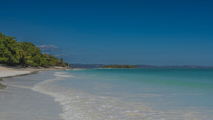 A beautiful tropical beach without people. The waves of the aquamarine ocean foam on the white sand. Green vegetation, palm trees on the shore. The blue sky. Copy space. Madagascar. 