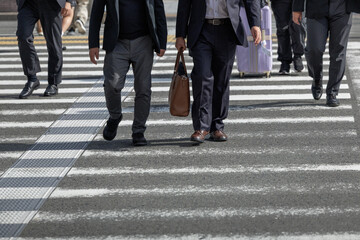 Obraz premium Several men in business attire cross city street via zebra crosswalk, walking among other casually dressed pedestrians. Scene captures urban movement and daily routine in a bustling environment.