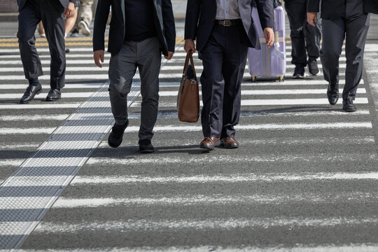 Several men in business attire cross city street via zebra crosswalk, walking among other casually dressed pedestrians. Scene captures urban movement and daily routine in a bustling environment.