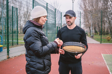 A tennis coach teaches a female student on an outdoor court, demonstrating proper technique with a ball and racket. Perfect for illustrating sports training, active lifestyle, or coaching sessions.