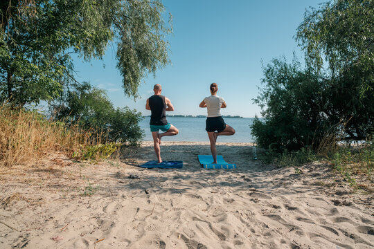 Couple practicing tree pose yoga on a serene sandy beach by the water under a clear blue sky. Perfect for wellness, mindfulness, and outdoor fitness themes. - Powered by Adobe