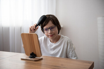 A boy in a white T-shirt combs his hair in front of a desk mirror standing on a wooden table in a softly lit interior.