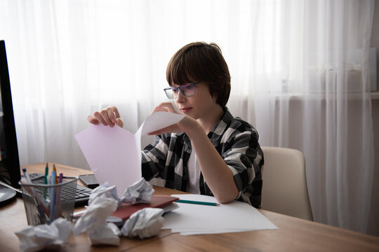 Young student at a desk crumpling paper while studying, surrounded by scattered sheets, stationery and a computer monitor.