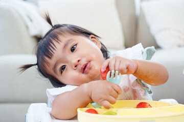 Asian toddler girl sitting highchair playing food pieces smiling joyfully home learning self feeding skill early development adorable baby enjoying mealtime happiness parenting family lifestyle indoor
