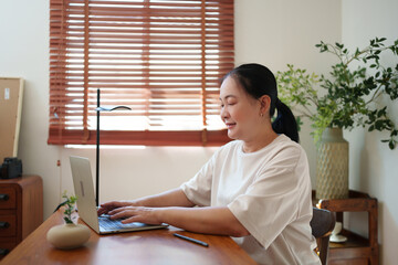Chinese middle-aged woman learning online from her laptop, focusing on her screen and enjoying a calm study moment at her wooden workspace.