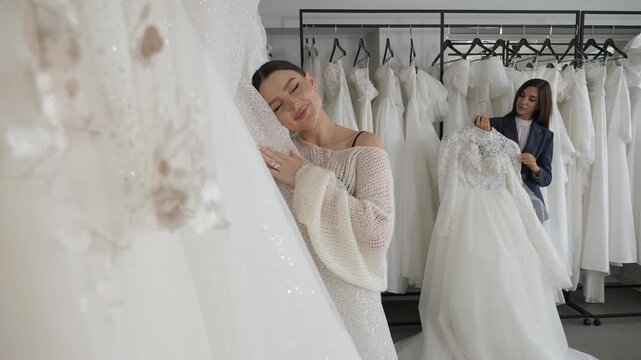 Two lovely brides are selecting a stunning wedding gown, standing in a spacious chamber filled with a plethora of gowns.