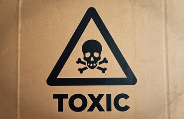 Black skull, crossbones symbol inside warning triangle on brown cardboard surface. Word TOXIC printed below. Hazard sign indicates dangerous chemicals biohazard risk. Warns about harmful substances.