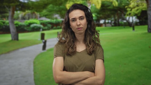 Woman in olive shirt crosses arms and looks sideways in a green park with a paved path under shaded trees; rejection.