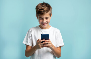 Fototapeta premium Young boy happily scrolls on smartphone. Teenager in white t-shirt smiles looking at phone screen. Isolated on light blue background. Child uses mobile device for fun, communication, or games online.