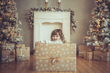 H&uuml;bsches kleines M&auml;dchen mit Locken sitzt in einem Geschenkkarton vor dem Weihnachtsbaum zu Weihnachten, Abendstimmung mit Lichtern Var. 5