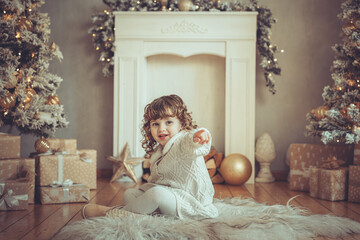H&uuml;bsches kleines M&auml;dchen mit Locken sitzt vor einem Kamin mit Weihnachtsbaum zu Weihnachten, Abendstimmung mit Lichtern Var. 3