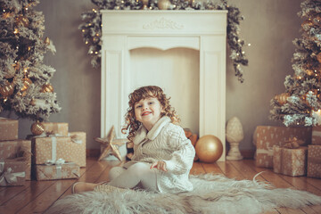 H&uuml;bsches kleines M&auml;dchen mit Locken sitzt vor einem Kamin mit Weihnachtsbaum zu Weihnachten, Abendstimmung mit Lichtern Var. 2
