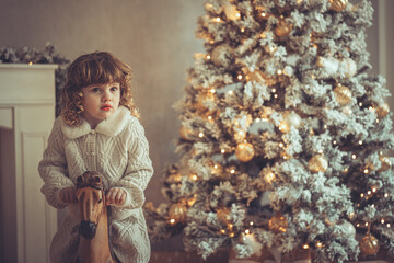 H&uuml;bsches kleines M&auml;dchen mit Locken sitzt auf einem Schaukelpferd neben einem Weihnachtsbaum zu Weihnachten, Abendstimmung mit Lichtern 