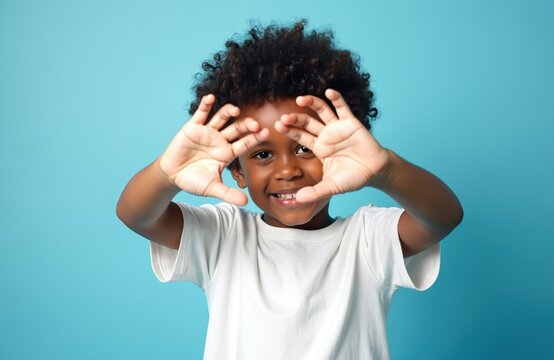 Cute african american child makes frame shape with hands. Adorable black boy looks at camera through fingers. Kid wears tshirt on blue background. Portrait of happy little child. - Powered by Adobe