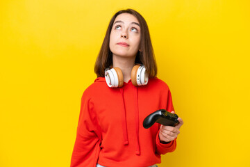 Young Ukrainian woman playing with a video game controller isolated on yellow background and looking up