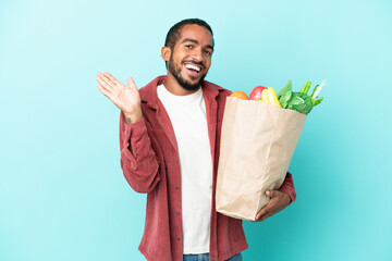 Young latin man holding a grocery shopping bag isolated on blue background saluting with hand with happy expression