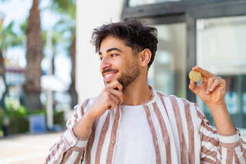 Handsome Arab man holding a Bitcoin at outdoors thinking an idea and looking side
