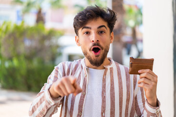 Handsome Arab man holding a wallet at outdoors surprised and pointing front