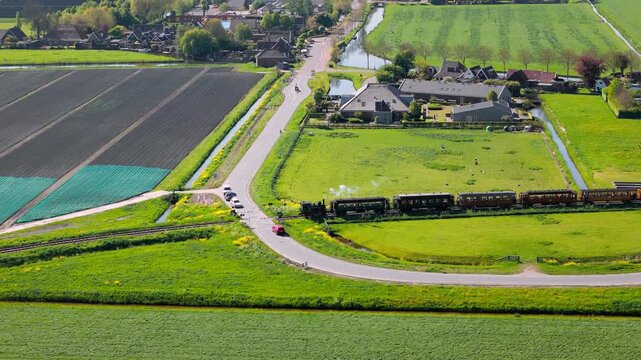 A vintage steam train crosses a quiet country road amidst vibrant green fields and farmlands. Traffic halts as the train passes through the peaceful Dutch landscape, evoking rural charm.