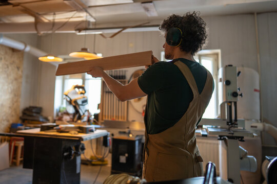 Careful carpenter man in earmuffs check examine wood plank before craft in carpentry studio. Quality control, woodworking occupation, craftsmanship, material inspection. Small furniture business.