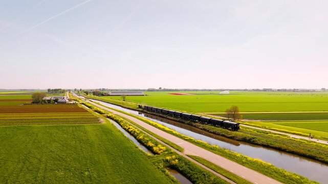 A nostalgic black steam train moves through colorful Dutch fields, passing canals and farms under a bright spring sky. The lush green landscape and vibrant colors evoke a peaceful rural mood. 