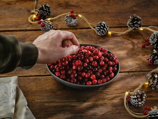A hand carefully selects vibrant cranberries from a bowl, surrounded by pinecones and twinkling lights. This scene captures the spirit of winter festivities and seasonal preparations