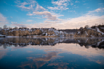 A serene winter landscape with a calm lake reflecting the cloudy sky and snow-covered cliffs.