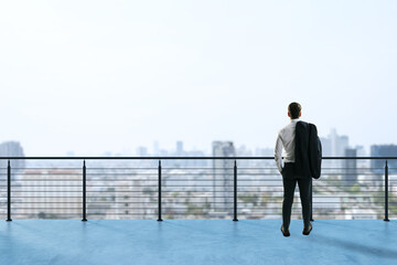 Obraz premium Businessman standing on rooftop terrace looking at city skyline in distance with a contemplative expression and jacket over shoulder.