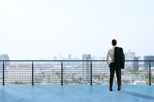 Businessman standing on rooftop terrace looking at city skyline in distance with a contemplative expression and jacket over shoulder.