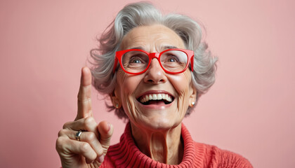 Joyful senior woman smiles widely, laughs, wears red glasses. Grey hair, points finger up. Elderly lady idea explanation on pink background, shows happy mood, energy, positive emotion.