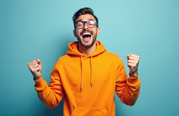 Fototapeta premium Young man with glasses shouts yes with joy. Pumps fists in triumph, excitement. Celebrates victory with happy, enthusiastic expression on face. Bright orange hoodie adds to vibrant look.