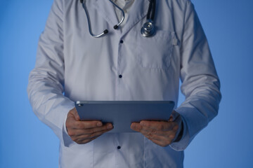 Doctor in white coat using digital tablet with stethoscope around neck against blue background, concept of modern healthcare and technology integration.