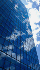 A modern blue glass skyscraper towers into a bright blue sky, its geometric patterns and reflective surfaces mirroring the sky and clouds.