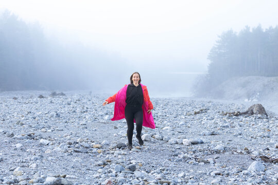 Woman in vibrant pink coat joyfully walks on rocky terrain in a misty landscape, surrounded by trees, capturing the essence of adventure and exploration in nature