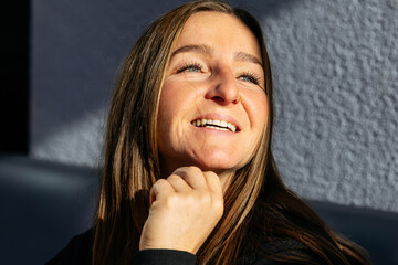 Smiling woman with long brown hair, illuminated by natural light, gazes thoughtfully while resting her chin on her hand, capturing a moment of joy and reflection