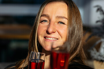 Young woman with long brown hair, smiling brightly while holding two glasses of red beverage, enjoying a joyful moment in a vibrant social setting with warm lighting