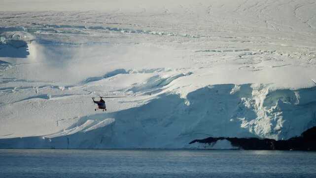 Red helicopter skims past towering ice cliffs along a frozen coastline, dwarfed by blue glacier walls and stratified snowfields above calm polar waters during an aerial traverse.