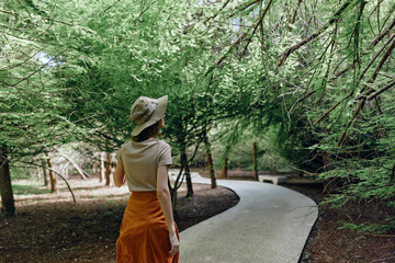 Woman walking on winding path through lush forest with hat and orange skirt, back view amid trees and nature trail, serene canopy light and peaceful outdoor leisure scene.