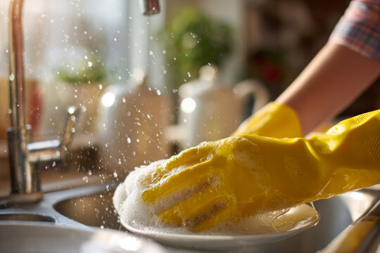 Person wearing yellow rubber gloves is washing dishes in a bright kitchen, with soap bubbles and water splashing, showcasing the cleaning process and home care activities - Powered by Adobe